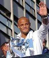 Vincent Kompany, de face, lors de la célébration de fin de saison 2013-2014. Il tient le trophée de la Coupe de la ligue d'Angleterre dans la main droite et fait un signe à la foule de la main gauche.